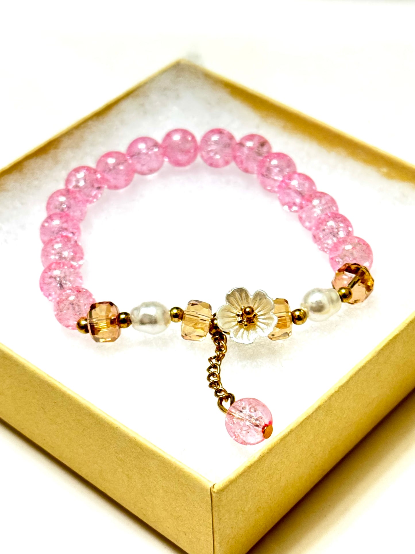 Handmade pink crackle beaded bracelet with white flower charm, gold crystals, and dangling bead, displayed in a gift box