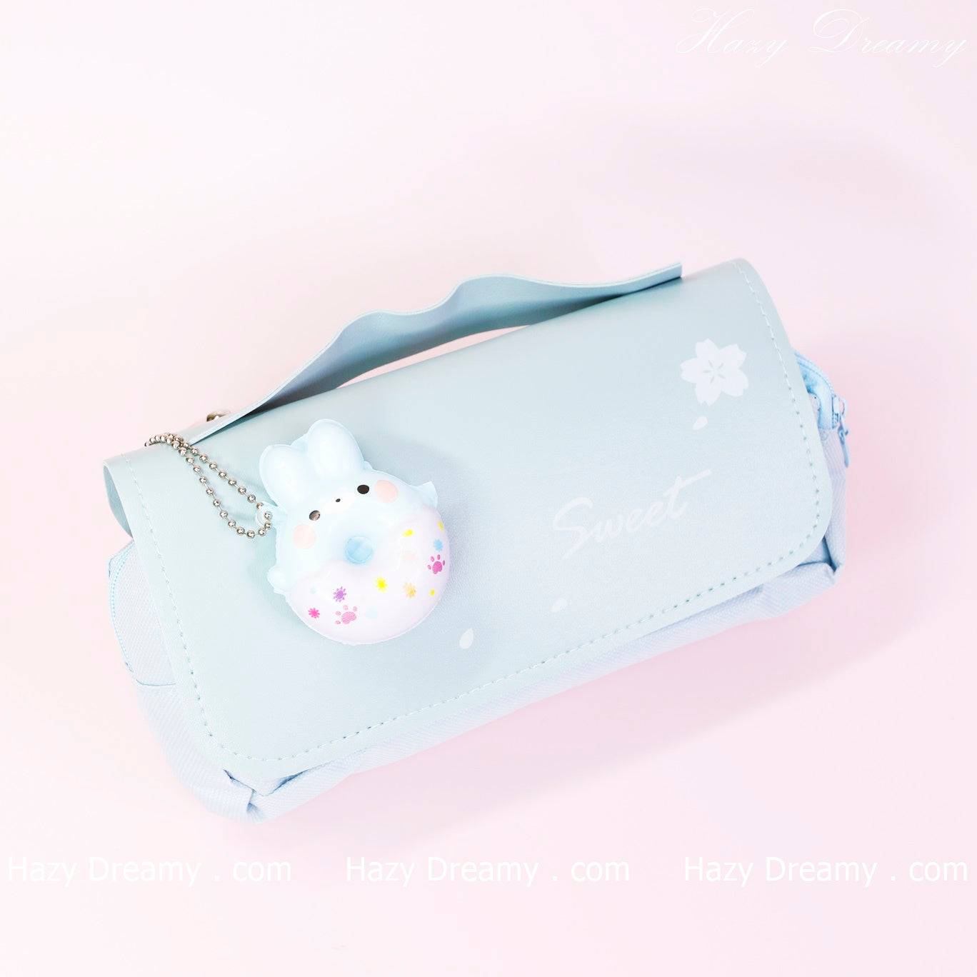 The image shows a light purple pencil case with a small, cute keychain attached to it. The keychain features a small, round character that resembles a bunny with a floral design. The word "Sweet" is written on the pencil case, which also has decorative elements like small flowers. The background of the image is also light purple, creating a soft and dreamy aesthetic. The brand name "Hazy Dreamy" is faintly visible in the top right corner
